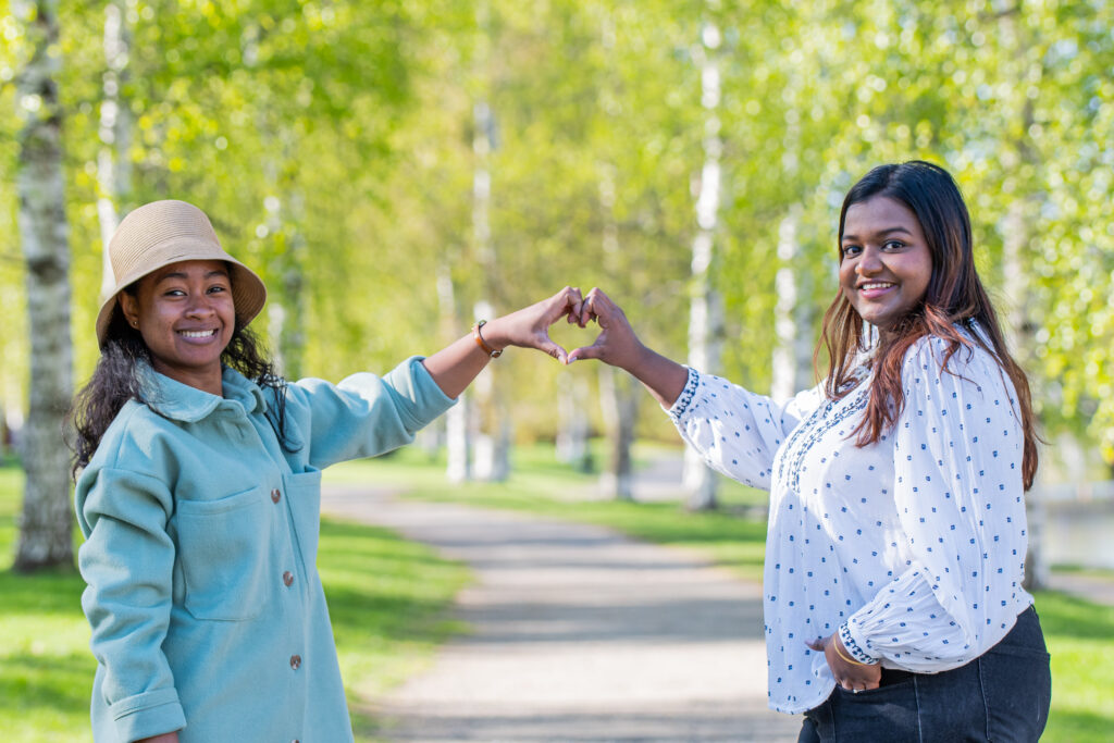 Two happy women in front of a sunny lane lined by birch trees form a heart with their hands and look towards the camera.